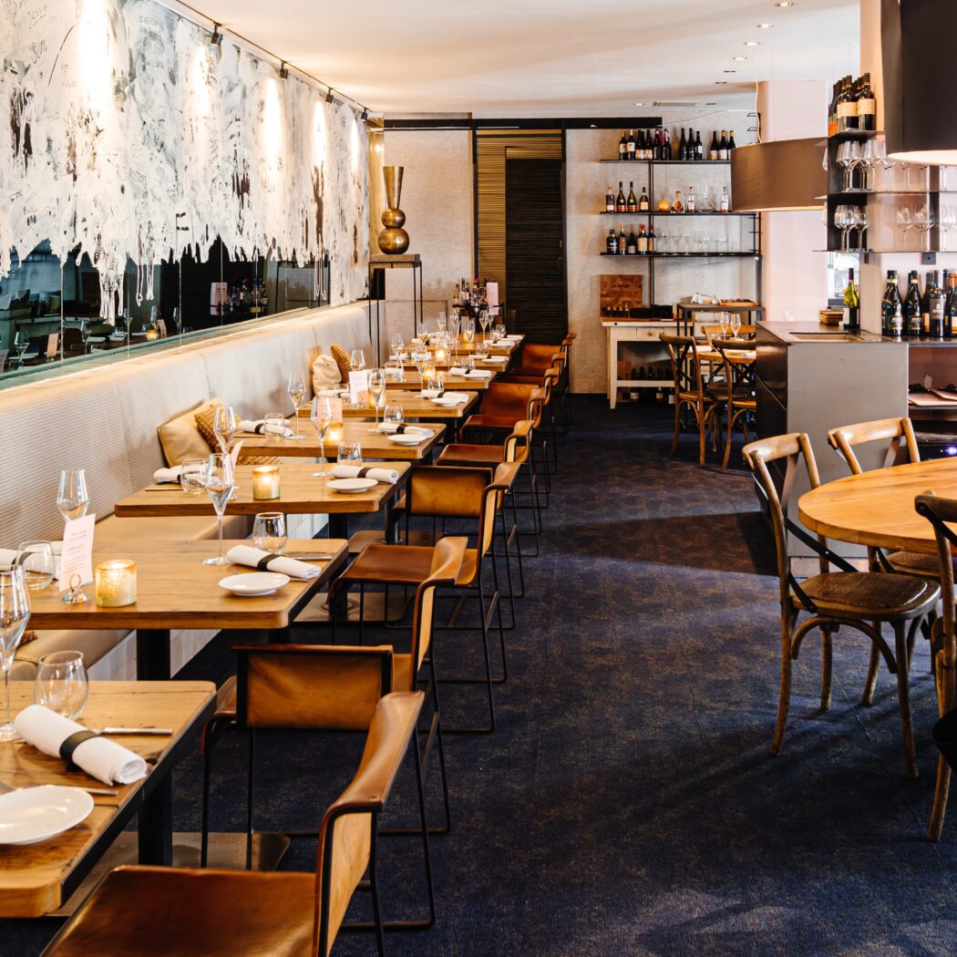 Modern restaurant met houten tafels, leren stoelen en een lange bank langs een spiegelwand met kunst.