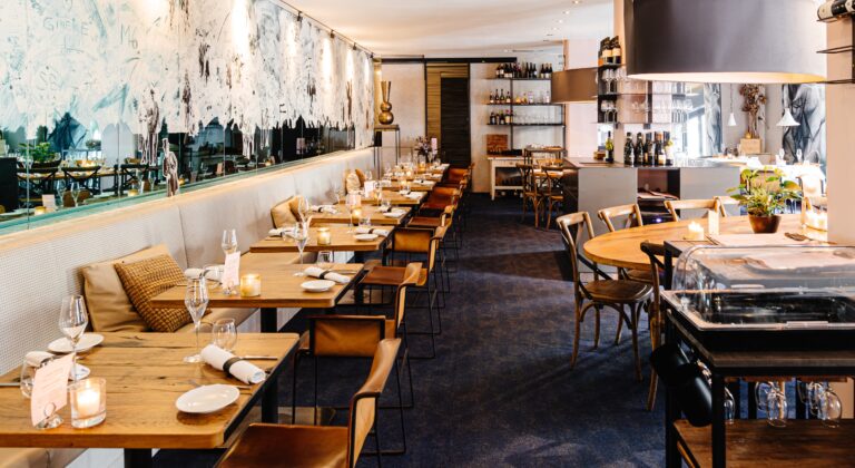 Modern restaurant met houten tafels, leren stoelen en een lange bank langs een spiegelwand met kunst.