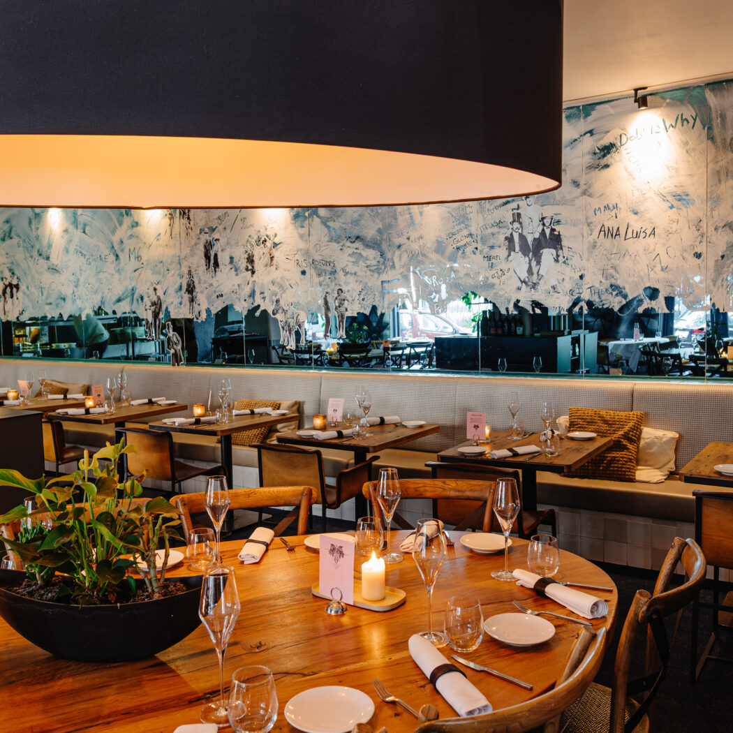 Sfeervol restaurant met een grote lampenkap, houten tafels, kaarsen, planten en abstracte muurkunst met ANA LUISA.