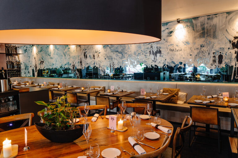 Sfeervol restaurant met een grote lampenkap, houten tafels, kaarsen, planten en abstracte muurkunst met ANA LUISA.