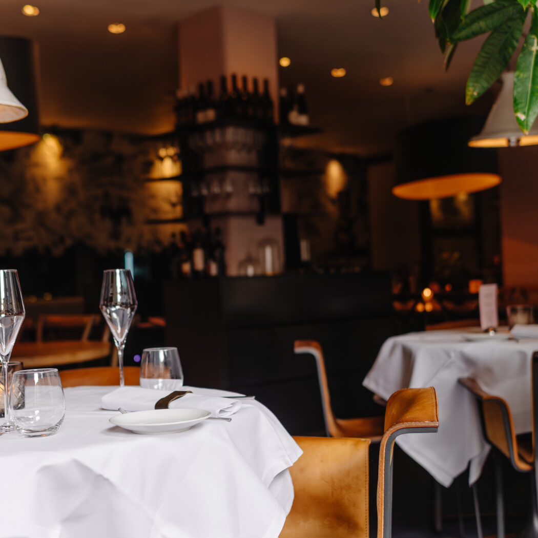 Gedekte restauranttafel met witte linnen, glazen, bestek en ‘DINNER MENU’ kaart, in warm verlicht interieur.