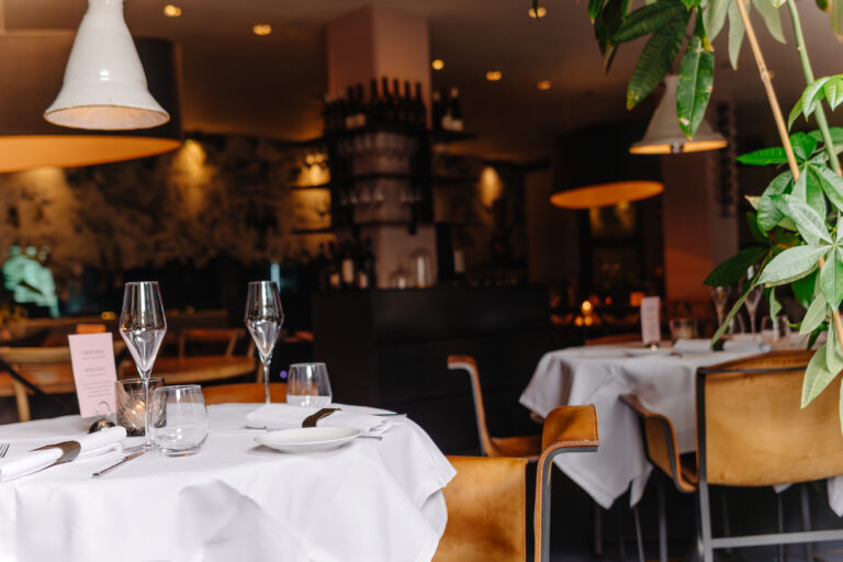 Gedekte restauranttafel met witte linnen, glazen, bestek en ‘DINNER MENU’ kaart, in warm verlicht interieur.
