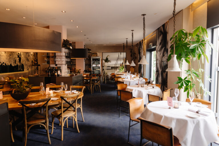 Sfeervol restaurant met houten en gedekte tafels, bruine stoelen, een bar en warme sfeerverlichting.