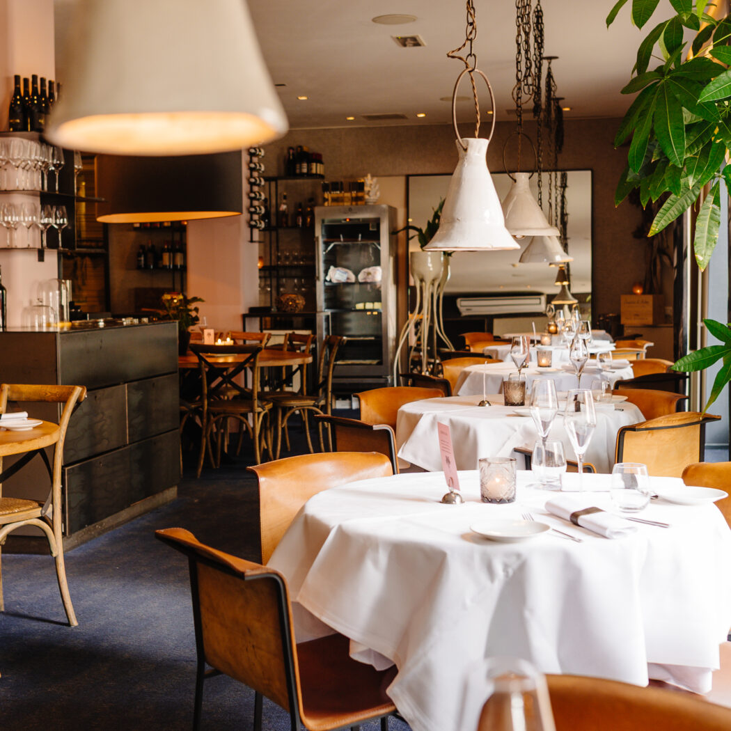 Sfeervol restaurant met gedekte tafels, bruine lederen stoelen, wijnbar en groene plant.