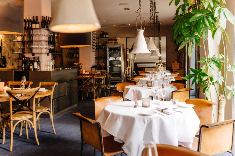 Sfeervol restaurant met gedekte tafels, bruine lederen stoelen, wijnbar en groene plant.