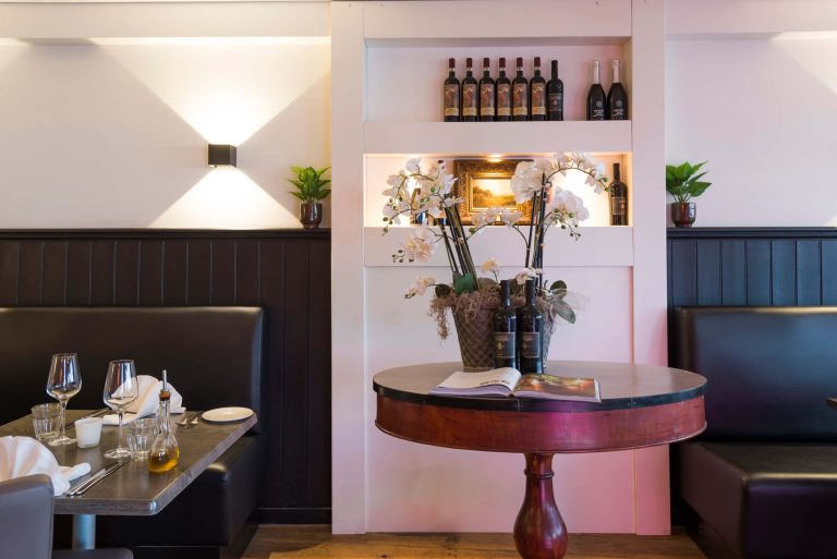 Sfeervol restaurantinterieur met gedekte tafels, donkere lederen banken, een bloemstuk van witte orchideeën en wijnflessen.