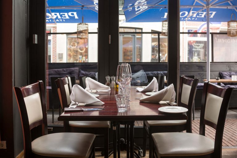 Netjes gedekte houten restauranttafel met glazen en witte servetten, met donkere stoelen en uitzicht op terras.