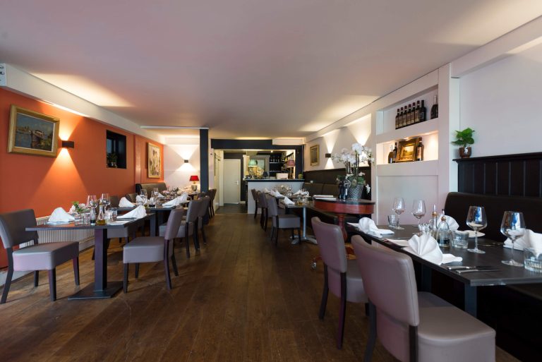 Warm verlicht restaurantinterieur met gedekte tafels, houten vloer, oranje muren en een bar op de achtergrond.
