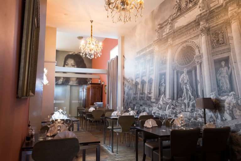 Restaurant met gedekte tafels, gouden kroonluchters en fotobehang van de Trevifontein en een vrouwengezicht.