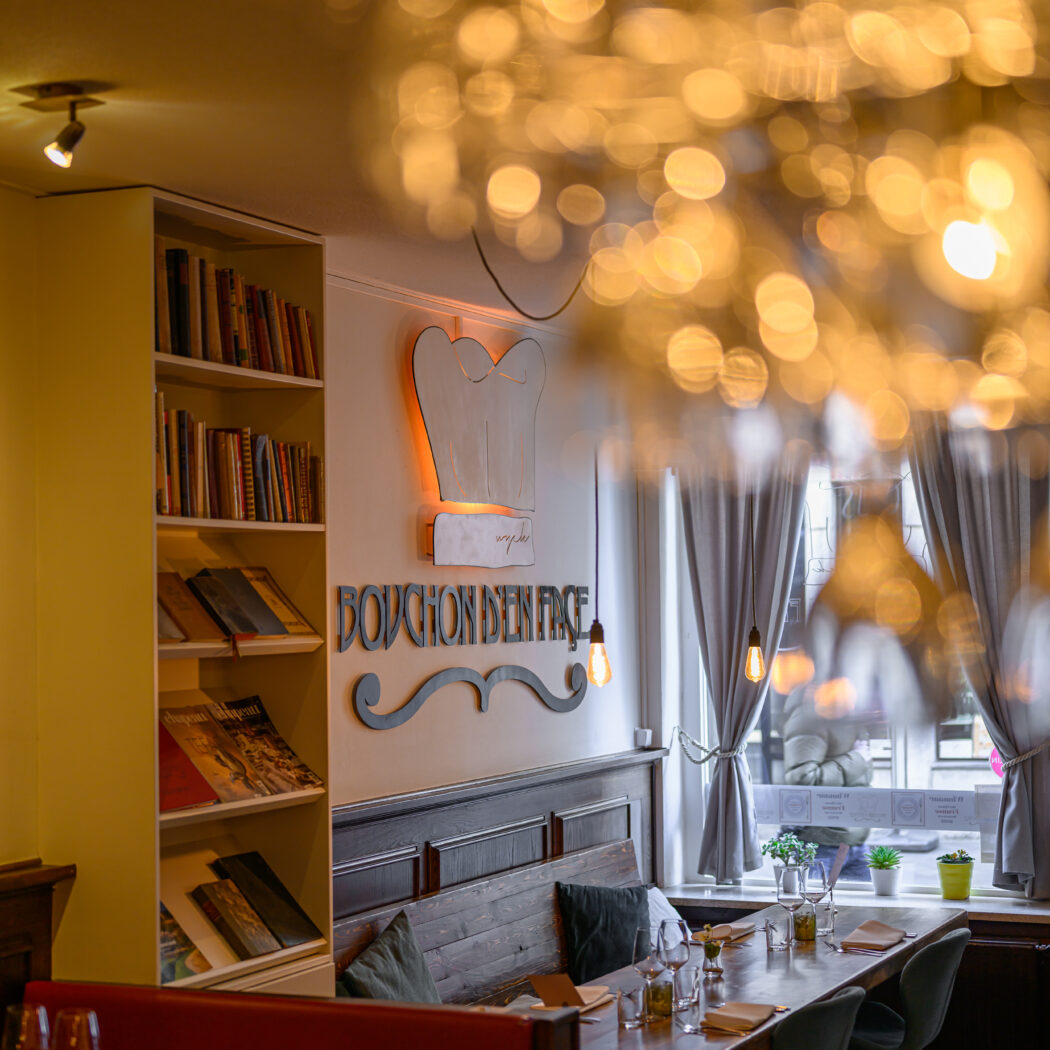 Sfeervol restaurantinterieur van BOUCHON DEN FACE met lange houten tafel, rode bank en verlichte kookmuts aan de muur.