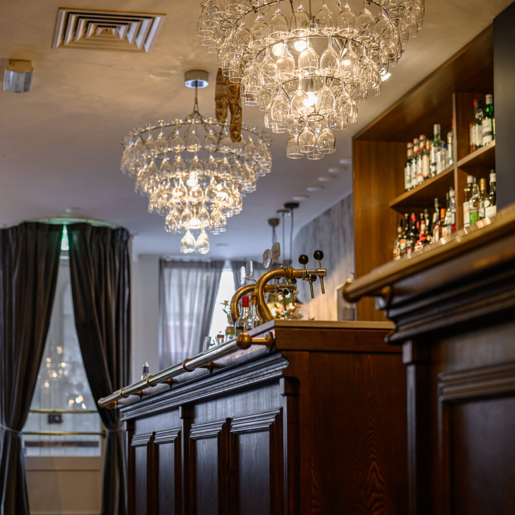 Sfeervol verlichte bar met donkerhouten toonbank, biertappen en kroonluchters van wijnglazen.