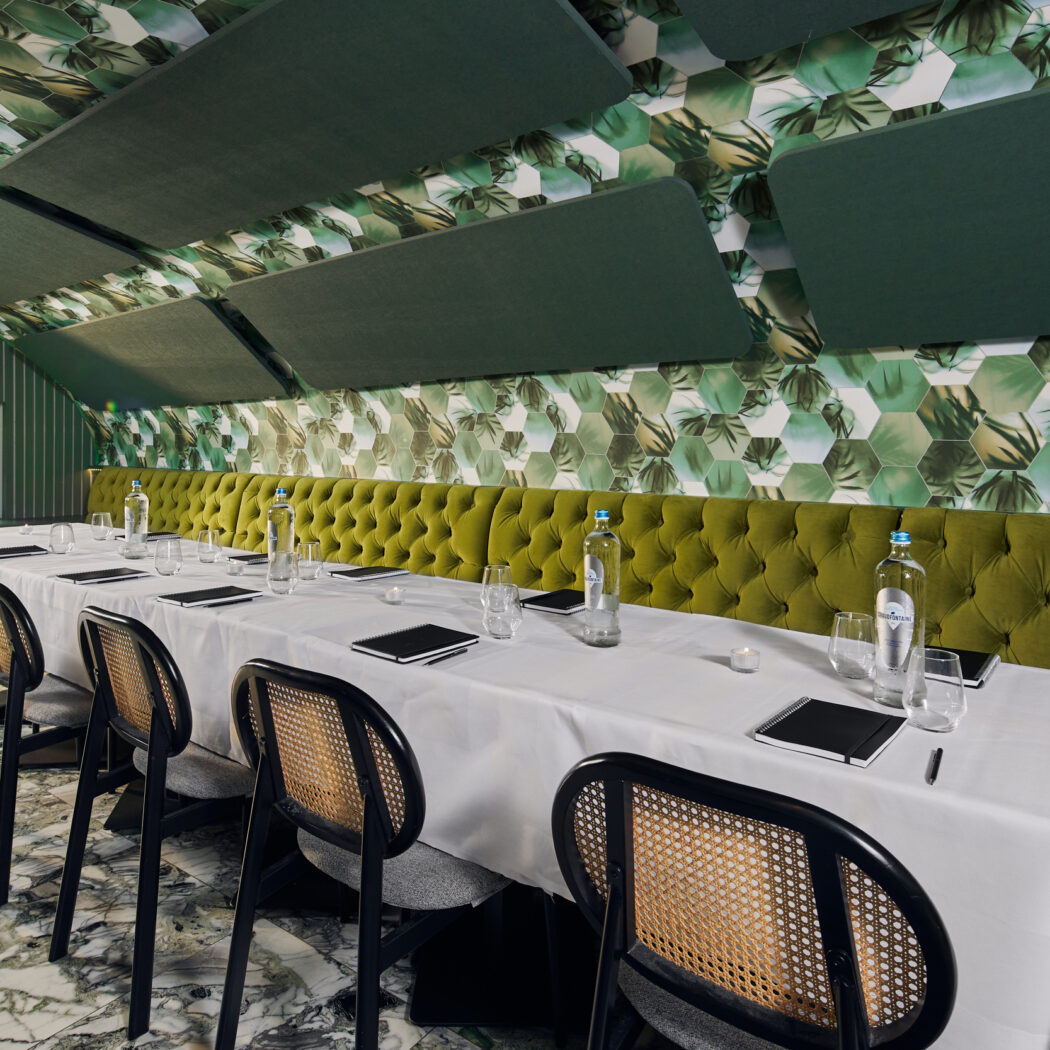 Stijlvolle vergaderruimte met lange witte tafel, groene bank, rieten stoelen en tropisch groen plafond.