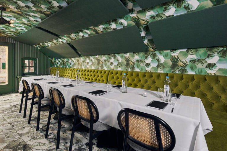 Stijlvolle vergaderruimte met lange witte tafel, groene bank, rieten stoelen en tropisch groen plafond.