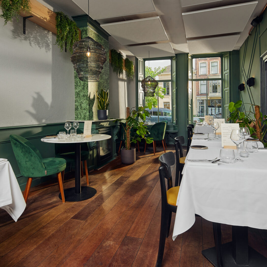 Sfeervol restaurant interieur met donkergroene muren, houten vloer, gedekte tafels, planten en kunstzinnige lampen.