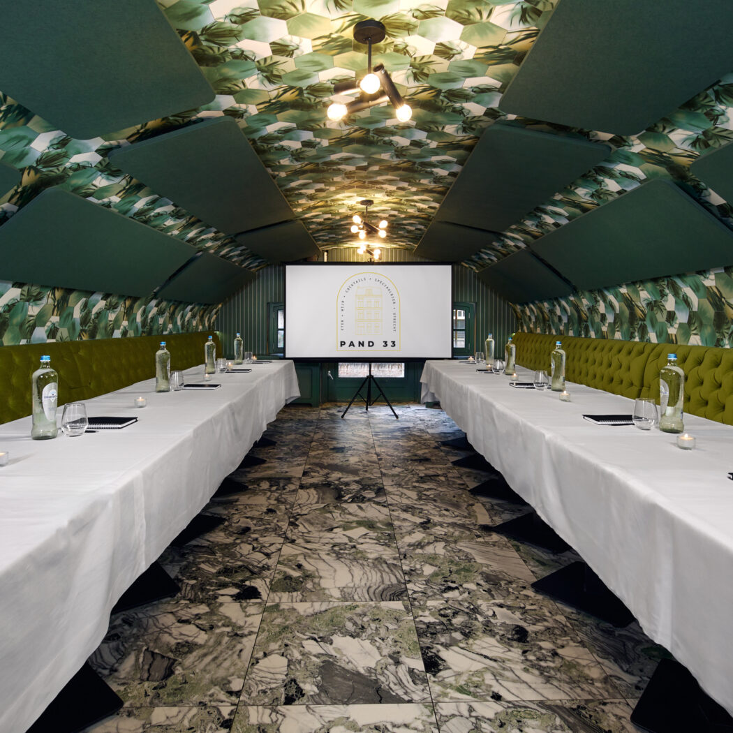 Sfeervolle vergaderzaal met wit gedekte tafels, groene banken, tropisch behang, marmeren vloer en presentatiescherm PAND 33.
