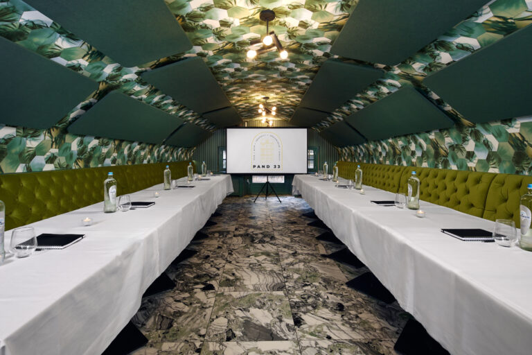 Sfeervolle vergaderzaal met wit gedekte tafels, groene banken, tropisch behang, marmeren vloer en presentatiescherm PAND 33.