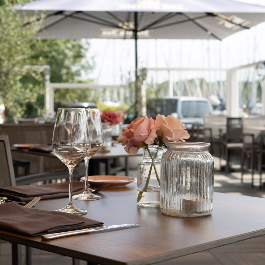 Gedekte restauranttafel op een zonnig buitenterras met wijnglazen, roze rozen en een theelichtje, aan de waterkant.