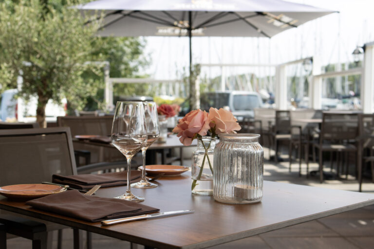 Gedekte restauranttafel op een zonnig buitenterras met wijnglazen, roze rozen en een theelichtje, aan de waterkant.