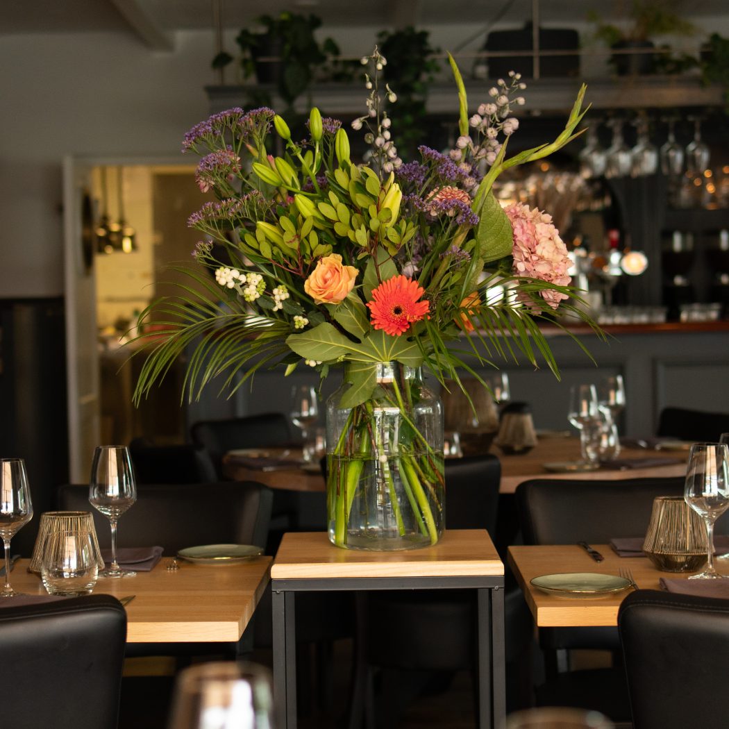 Kleurrijk bloemstuk met oranje gerbera en roos in glazen vaas op een houten tafeltje in restaurant.
