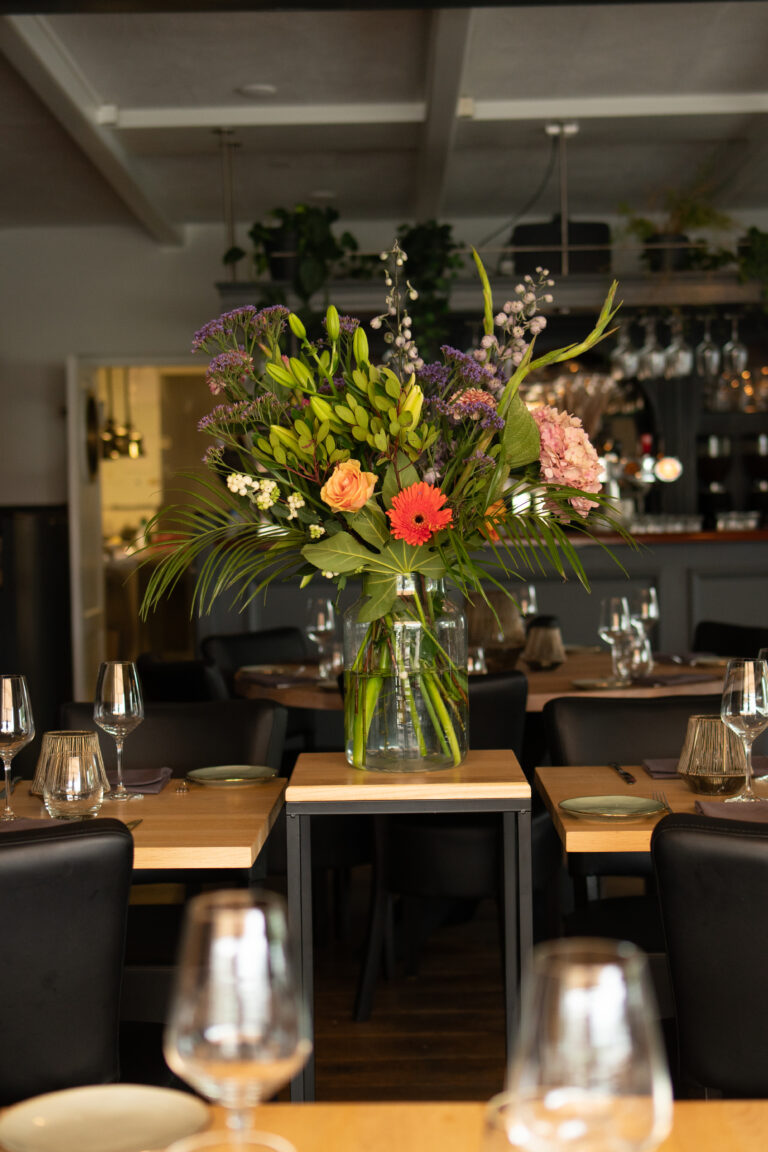 Kleurrijk bloemstuk met oranje gerbera en roos in glazen vaas op een houten tafeltje in restaurant.