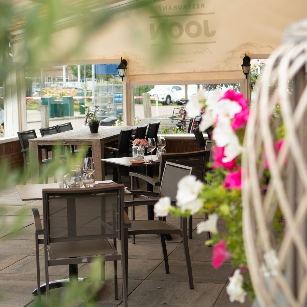 Gedeeltelijk overdekt restaurantterras van Restaurant Loon met houten tafels, stoelen en roze-witte bloemen.