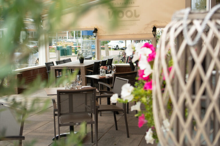 Gedeeltelijk overdekt restaurantterras van Restaurant Loon met houten tafels, stoelen en roze-witte bloemen.