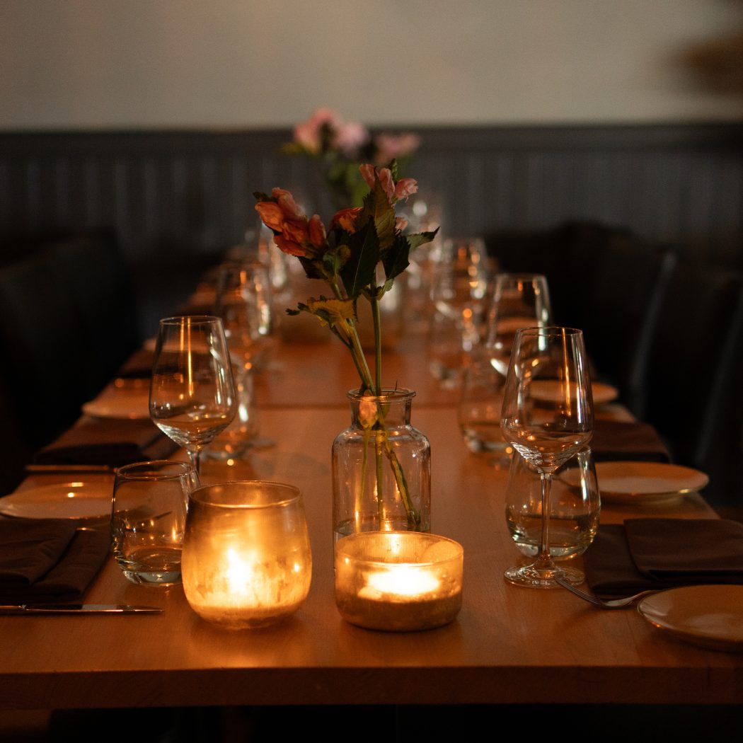 Intiem gedekte houten restauranttafel met wijnglazen, borden, bloemen en warm kaarslicht.