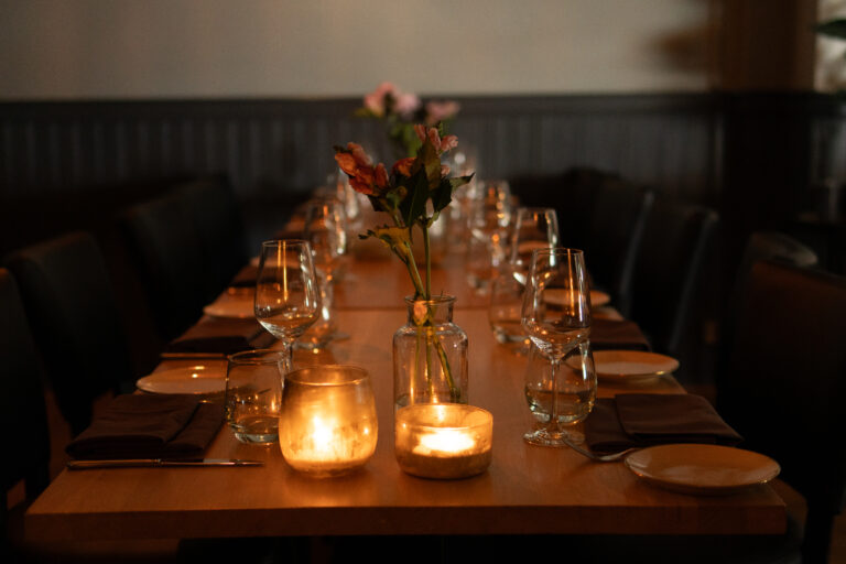 Intiem gedekte houten restauranttafel met wijnglazen, borden, bloemen en warm kaarslicht.