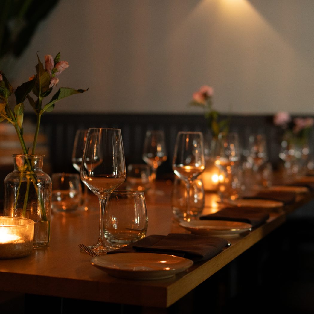 Houten eettafel gedekt met wijnglazen, kaarslicht en roze bloemen voor een warm diner.