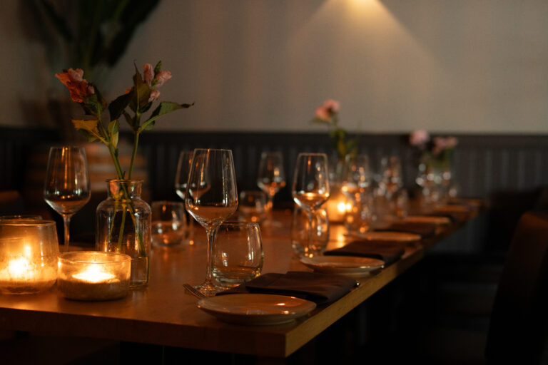 Houten eettafel gedekt met wijnglazen, kaarslicht en roze bloemen voor een warm diner.