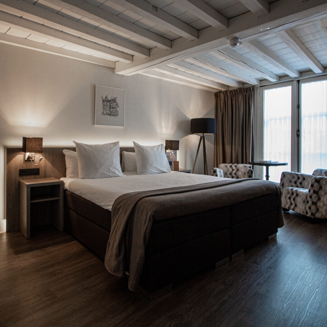 Sfeervolle hotelkamer met comfortabel tweepersoons boxspringbed, twee fauteuils, houten vloer en wit balkenplafond.