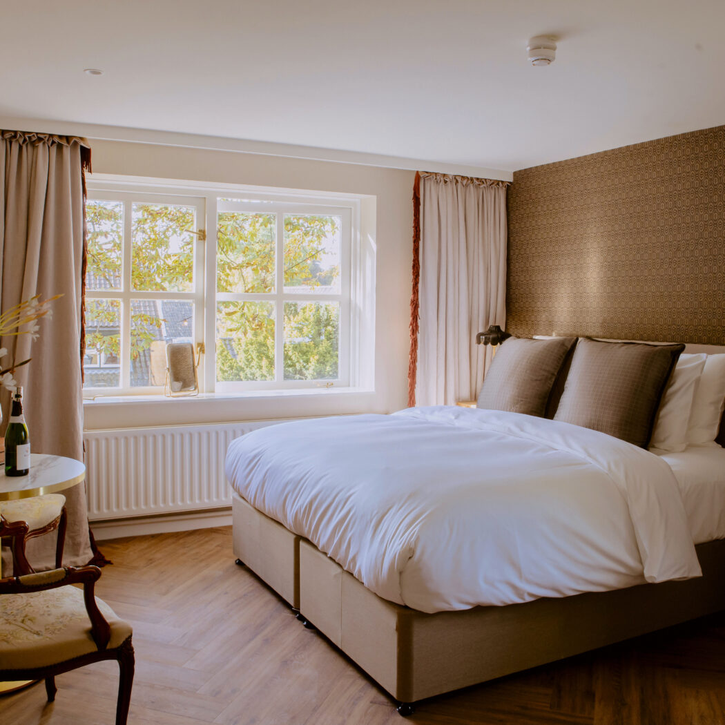 Sfeervolle hotelkamer met tweepersoonsbed, donkerbruin behang, raam met groene bomen en fles champagne.