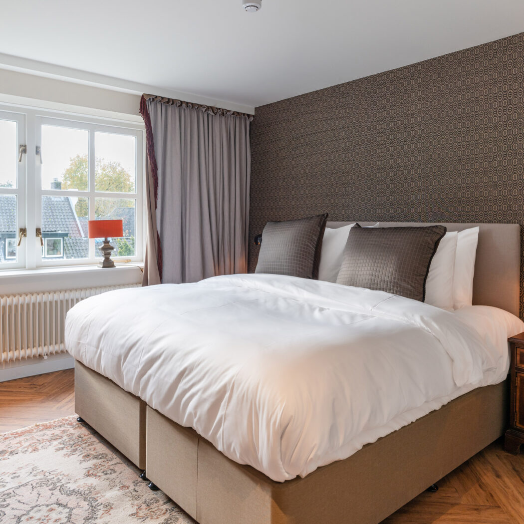 Opgemaakt tweepersoonsbed in sfeervolle slaapkamer met donkerbruin patroonbehang, houten visgraatvloer, beige gordijnen en tafellampen.