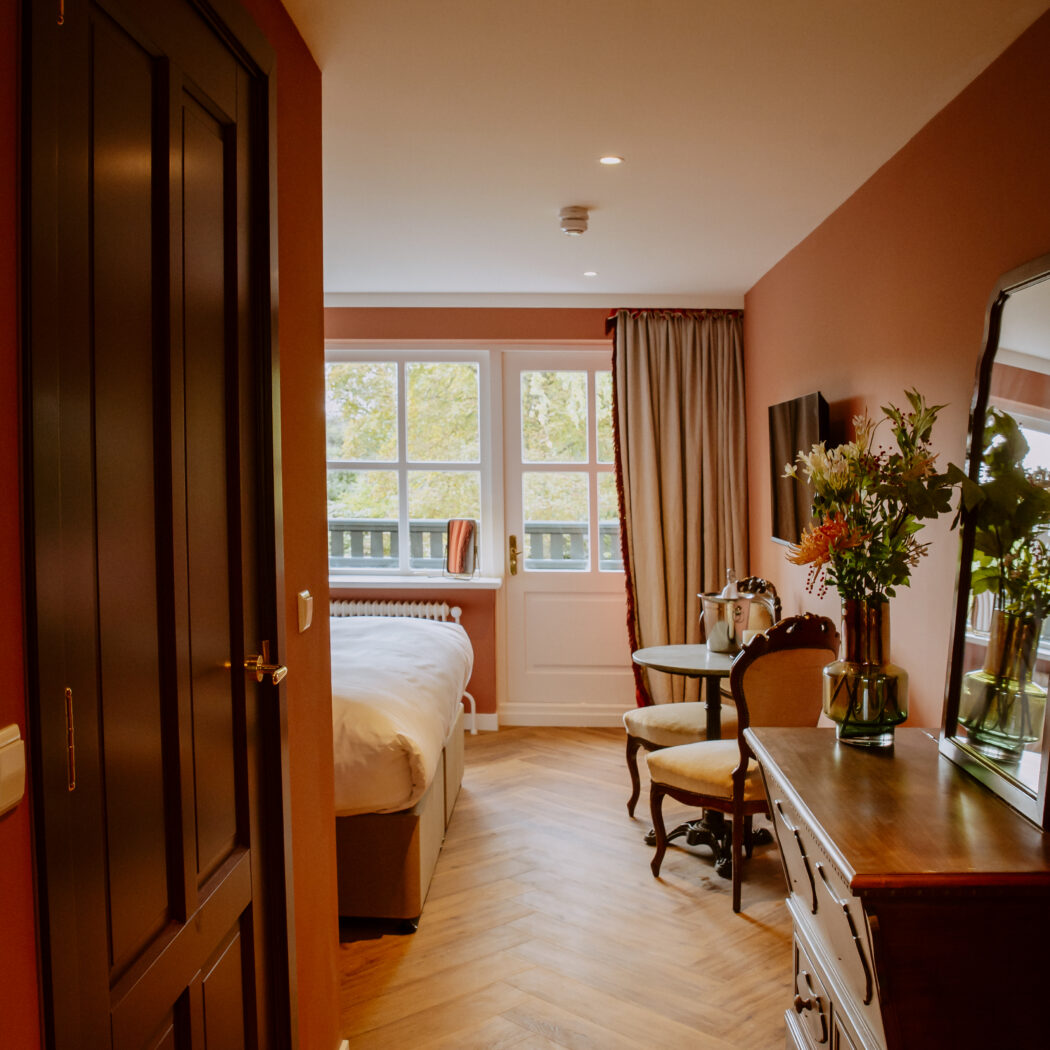 Een warme hotelkamer met open donkere deur, terracotta muren, visgraatvloer, bed, balkon en houten dressoir met bloemen.
