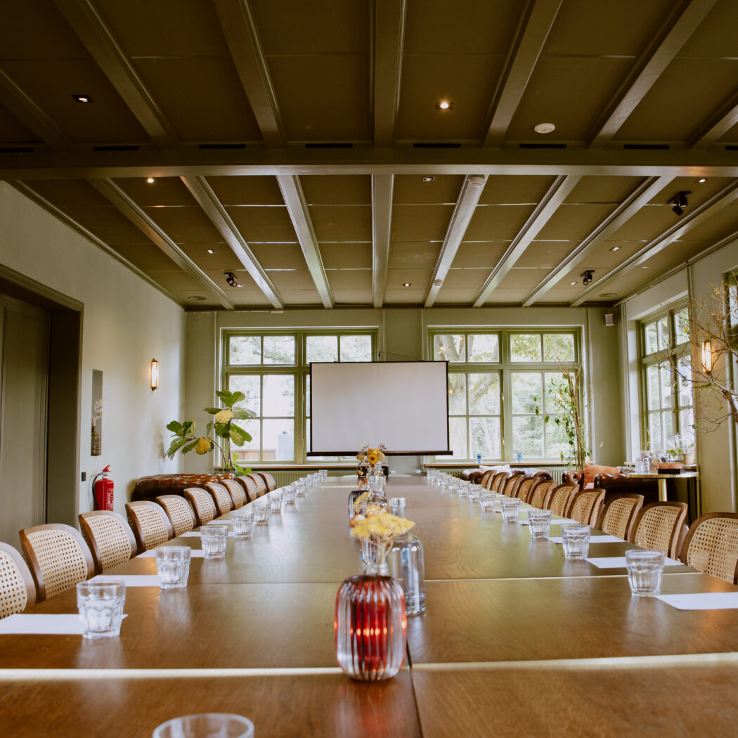 Sfeervolle vergaderzaal met lange houten tafel, rieten stoelen, projectiescherm en grote ramen met groen uitzicht.