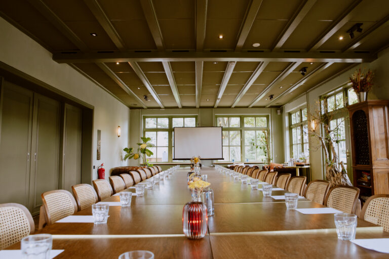 Sfeervolle vergaderzaal met lange houten tafel, rieten stoelen, projectiescherm en grote ramen met groen uitzicht.