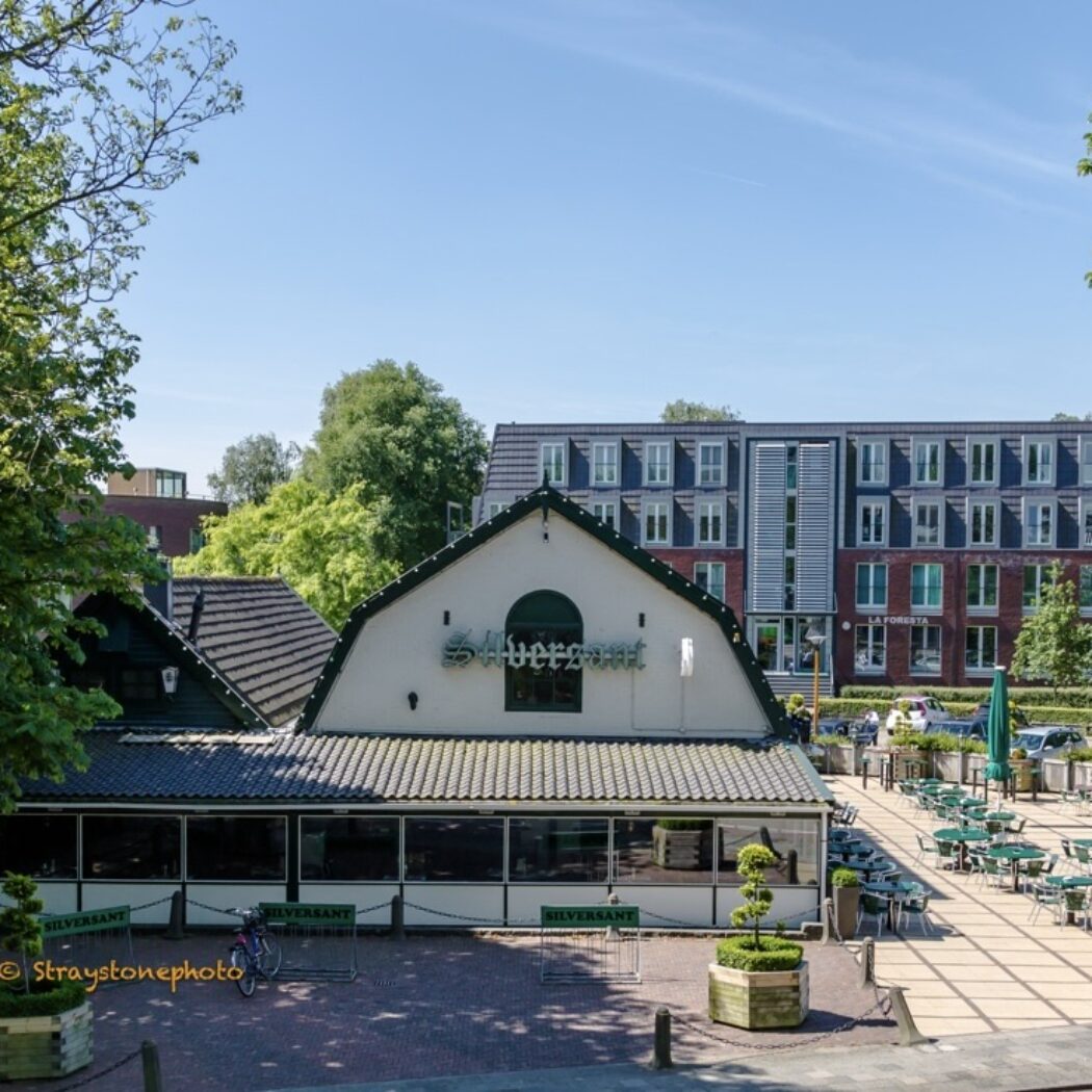Cafe-restaurant Silversant met een groot buitenterras vol groene stoelen en tafels op een zonnige dag, naast appartementencomplex La Foresta.