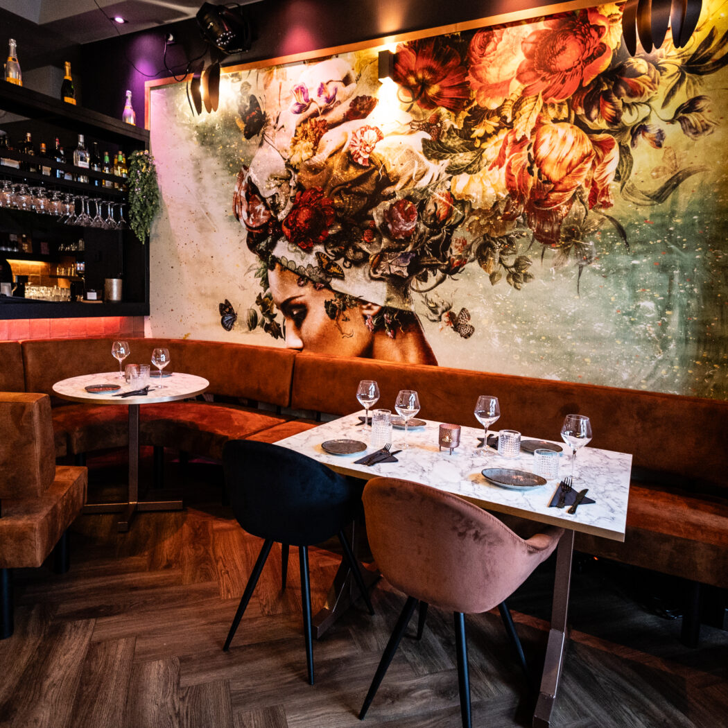Modern restaurant met opvallende muurschildering van vrouw met bloemen en vlinders, bruine zitbank en gedekte tafels.