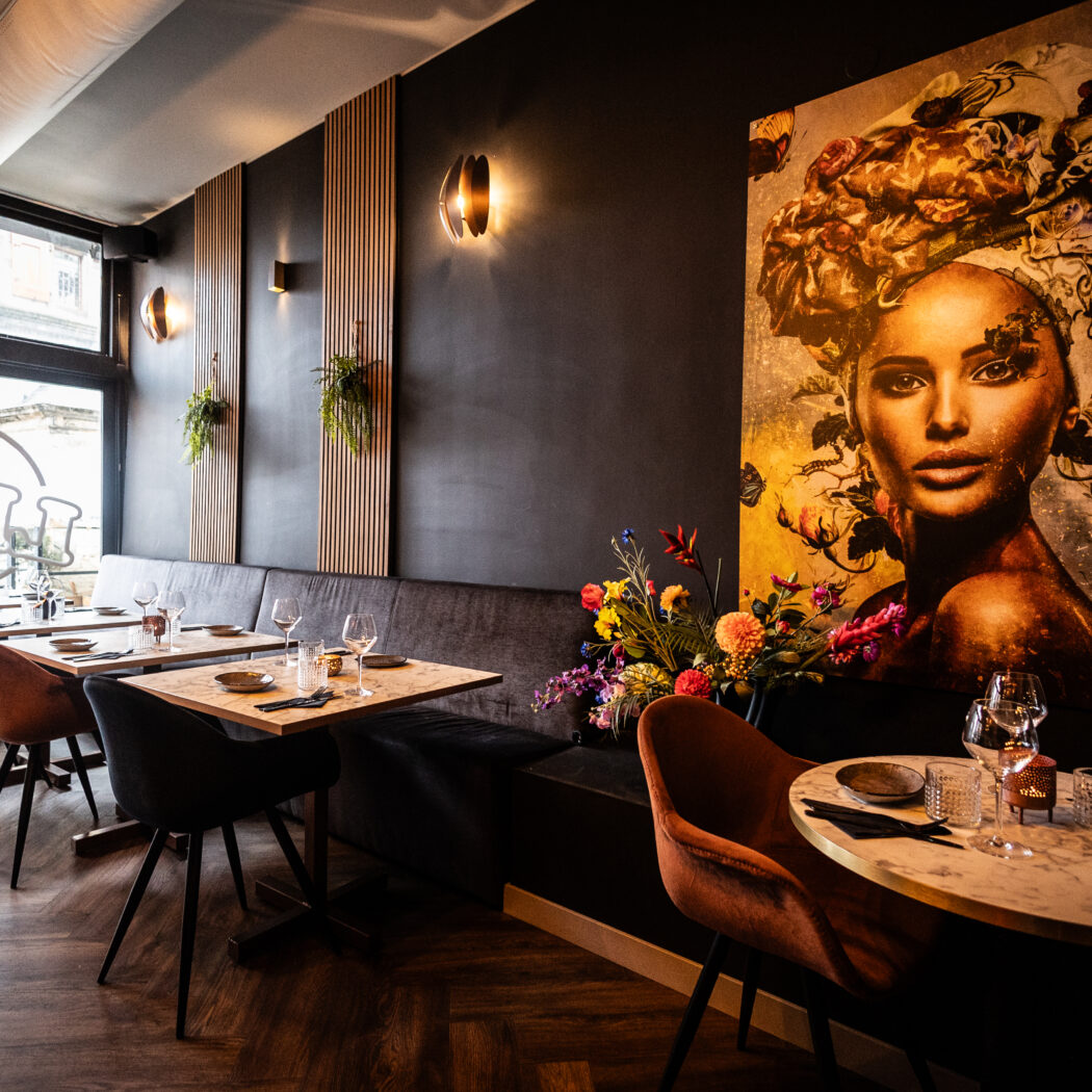 Sfeervol restaurant met donkere muren, houten lamellen, bloemig kunstwerk, marmeren tafels en BAK op het raam.