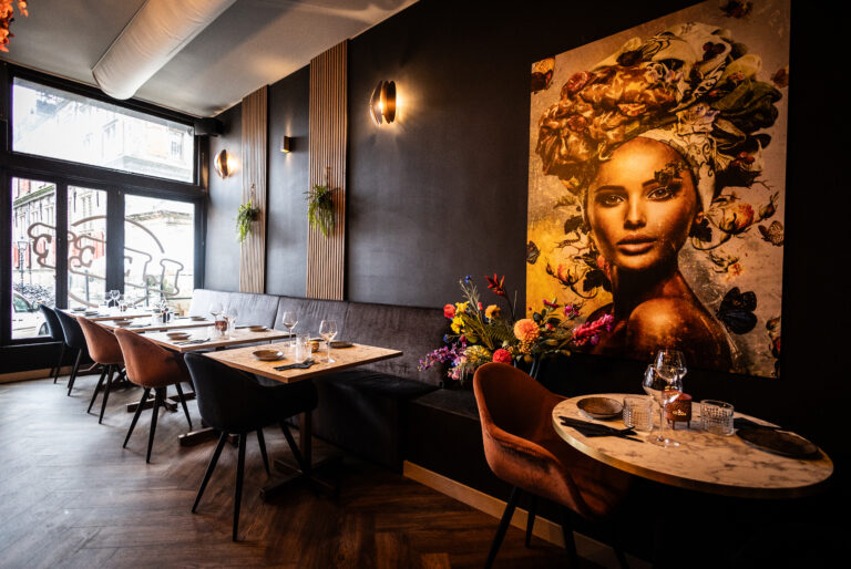Sfeervol restaurant met donkere muren, houten lamellen, bloemig kunstwerk, marmeren tafels en BAK op het raam.