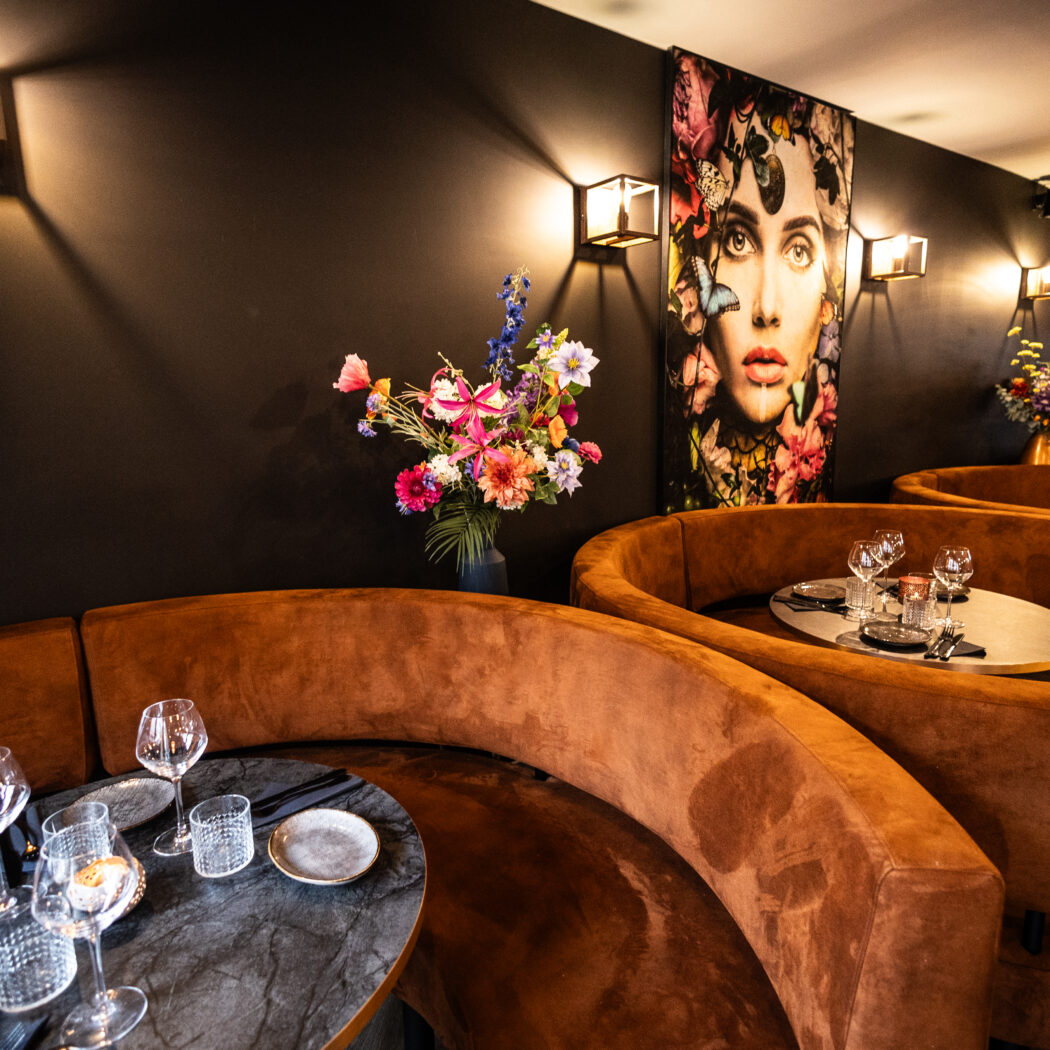Sfeervol restaurantinterieur met gebogen bruine fluwelen banken, gedekte ovale tafels en een groot bloemrijk kunstwerk.