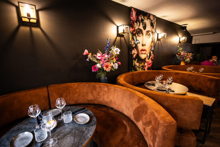 Sfeervol restaurantinterieur met gebogen bruine fluwelen banken, gedekte ovale tafels en een groot bloemrijk kunstwerk.