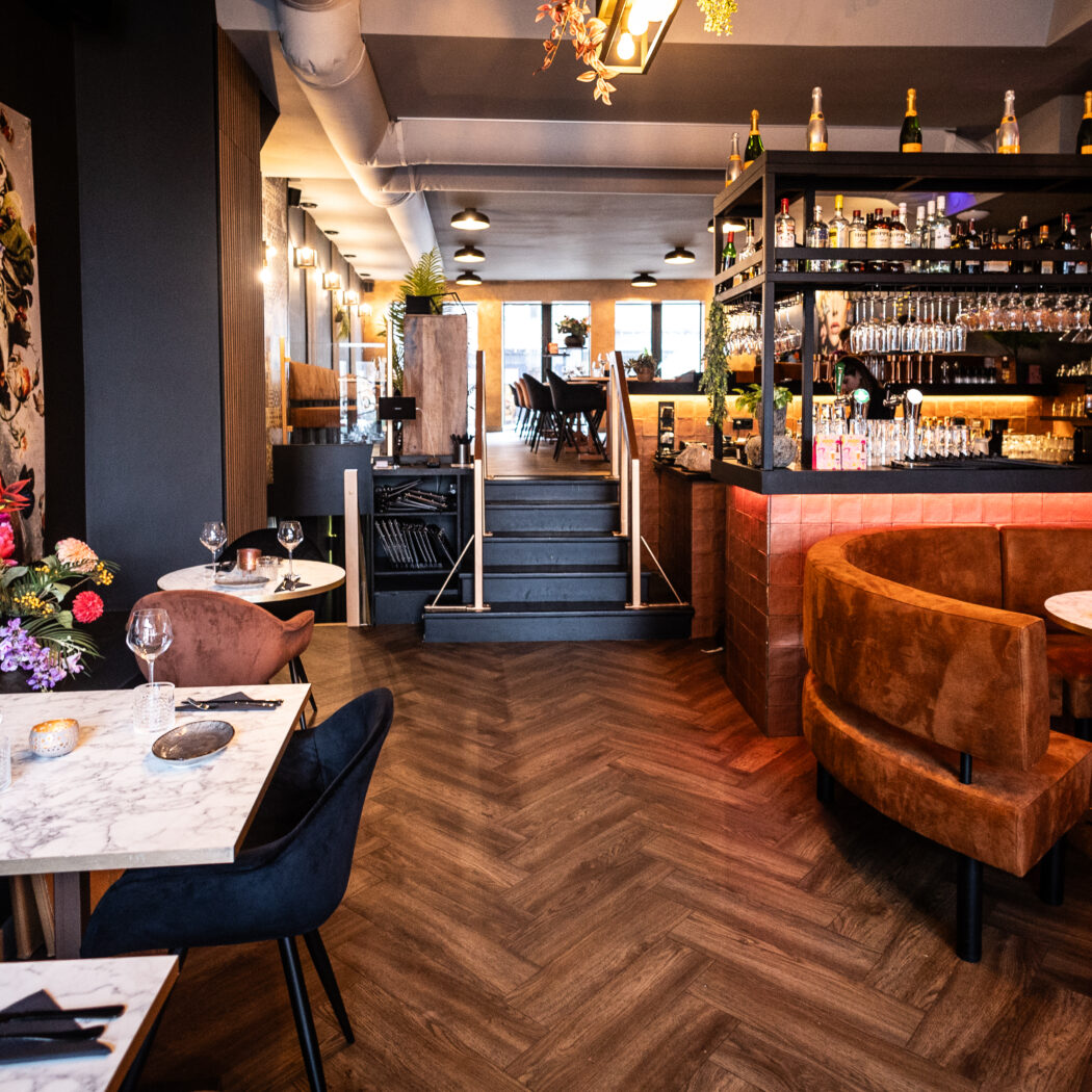 Modern, chique restaurant met verlichte bar, ronde bruine fluwelen banken, marmeren tafels en kleurrijk kunstwerk.