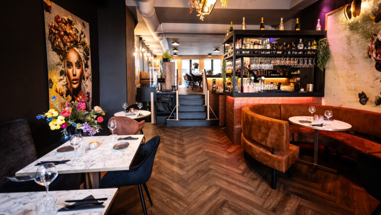 Modern, chique restaurant met verlichte bar, ronde bruine fluwelen banken, marmeren tafels en kleurrijk kunstwerk.
