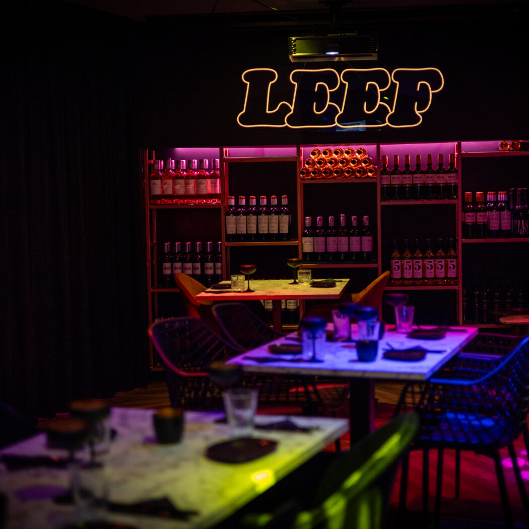 Sfeervol restaurant met roze verlichte flessenrekken, gedekte tafels en een oplichtend LEEF neonteken.