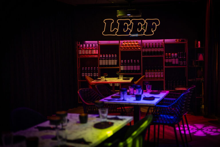 Sfeervol restaurant met roze verlichte flessenrekken, gedekte tafels en een oplichtend LEEF neonteken.