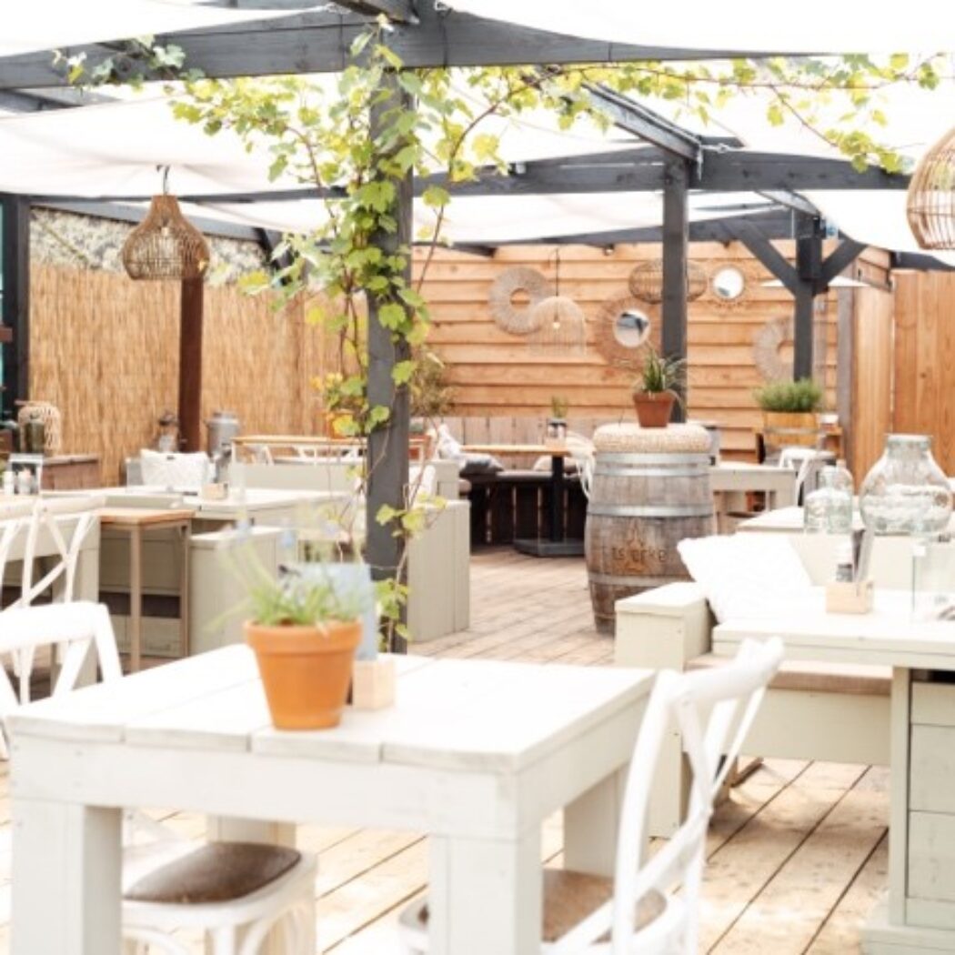 Gezellig restaurantterras met witte houten meubels, rieten lampen en een pergola met groene klimplanten.