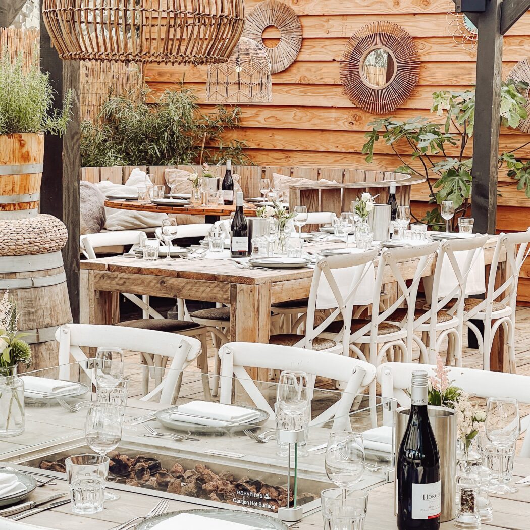 Buitenterras met gedekte houten tafels, witte stoelen, rotan lampen en een tafel met easyfires vuurpit.