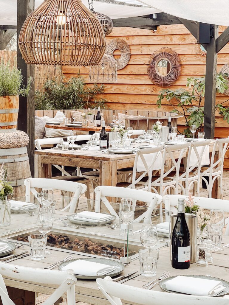 Buitenterras met gedekte houten tafels, witte stoelen, rotan lampen en een tafel met easyfires vuurpit.