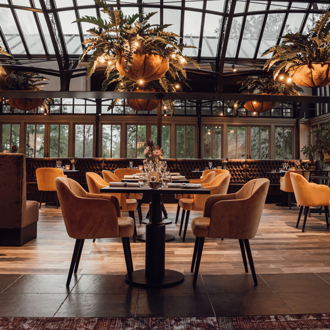 Sfeervolle restaurantserre met glazen dak, gedekte tafels, oranje fluwelen stoelen en verlichte hangplanten.